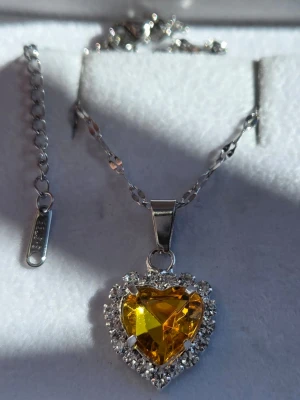 Halsband med hjärta och gul sten - Snyggt halsband i silverfärg med ett hjärtformat hänge. Hänget har en stor gul, glittrande sten i mitten och är omgiven av små klara stenar som ger extra bling. Kedjan är tunn i rostfritt stål och har ett justerbart lås. Perfekt för dig som gillar att sticka ut med accessoarer.