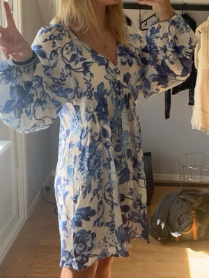 Blommig kortklänning med ballongärm - Supersöt kortklänning med blått blommönster på vit botten. Klänningen har v-ringning och vida ballongärmar med resår vid handlederna. Perfekt för sommarens alla tillfällen och ger en fräsch och romantisk vibe.