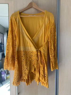 Gul omlottblus med spetsdetaljer från Odd Molly - Säljer en kort gul omlottblus med v-ringning och knyt i midjan. Blusen har långa ärmar och vackra spetsdetaljer samt volangkant nedtill. Perfekt för sommarens alla tillfällen och ger en bohemisk vibe.