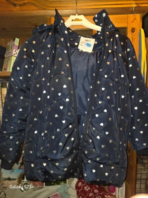 Mörkblå pufferjacka med hjärtmönster - Snygg mörkblå pufferjacka från Ellos med silverfärgade hjärtan över hela jackan. Jackan har huva, dragkedja framtill och två stora fickor. Materialet är glansigt och quiltat för extra värme. Perfekt för kalla vinterdagar.