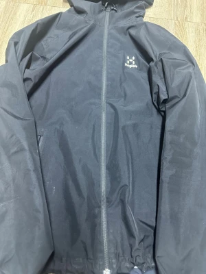 Svart regnjacka från Haglöfs - Snygg svart regnjacka från Haglöfs med hel dragkedja framtill och justerbar huva. Jackan har diskret logga på bröstet och meshfoder på insidan för extra ventilation. Perfekt för regniga dagar och outdoor-äventyr.