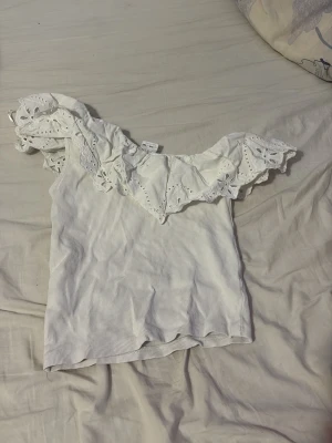 Vit offshoulder topp med spetsvolang - Säljer en söt vit offshoulder topp med bred spetsvolang och ribbad struktur. Toppen är ärmlös och har fina broderade detaljer i volangen som ger en romantisk vibe. Perfekt för varma dagar och somriga outfits. Storlek 11-12 men passar mig som har xs!