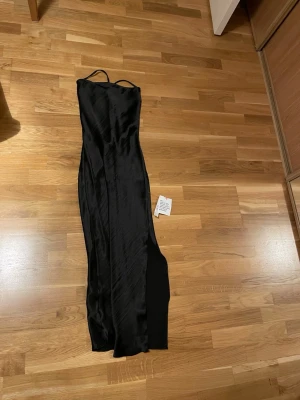 Svart långklänning med öppen rygg - Snygg svart långklänning i satinliknande material med tunna axelband och öppen rygg med snörning. Klänningen har en elegant, figurnära passform och slits på sidan. Perfekt för dig som vill sticka ut på nästa event.