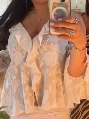 Vit volangblus Zara - vit blus med blommigt mönster och volangdetaljer längs ringningen och framtill. Tillverkad i ett lätt, transparent tyg som ger en luftig känsla. Blusen har långa ärmar och en v-ringning. Perfekt för dig som gillar en söt och trendig stil. Storlek S/M