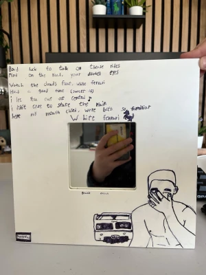 Spegel med Frank Ocean-tema - Unik spegel med handritad Frank Ocean-inspirerad design. Vit ram med svart tuschteckning av Frank Ocean, kassettband och handskrivna låttexter från 'White Ferrari'. Spegeln är fyrkantig och ramen är i hård kartong eller trä. Ikea malma spegel ny kostar ungefär 200kr