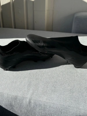 Svarta Adidas fotbollsskor utan snörning - Säljer ett par helsvarta fotbollsskor från Adidas med klassiska tre ränder på sidan. Skorna har en strömlinjeformad design utan snörning och är tillverkade i syntetmaterial med textildetaljer. Perfekta för dig som vill ha stilrena och snabba skor på planen.
