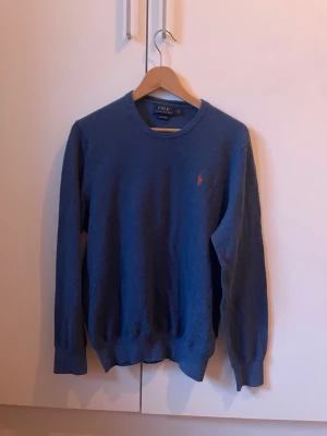 Blå tröja från Polo Ralph Lauren - Klassisk blå tröja från Polo Ralph Lauren med rund halsringning och den ikoniska röda logotypen broderad på bröstet. Tröjan har långa ärmar och ribbade muddar vid ärmslut och nederkant. Perfekt för en stilren och avslappnad look.