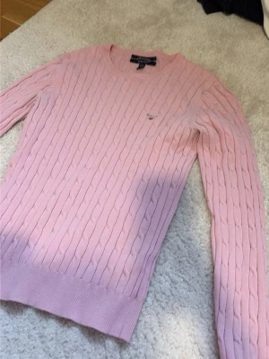 Ljusrosa kabelstickad tröja Gant - Supersnygg ljusrosa kabelstickad tröja från Gant med rund halsringning och diskret logga broderad på bröstet. Tröjan har långa ärmar och ribbade muddar vid ärmslut och nederkant. Perfekt för en klassisk och fräsch look.