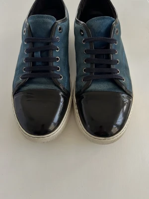 Blå sneakers från Lanvin med lackad tå - Snygga blå sneakers från Lanvin med svart lackad tå och vita sulor. Ovandelen är i mocka och skorna har svarta skosnören. Klassisk låg modell som passar till många olika stilar. Inkluderar box. Skorna är 9/10 och lacken är 10/10. Passar strl 42 och 43.