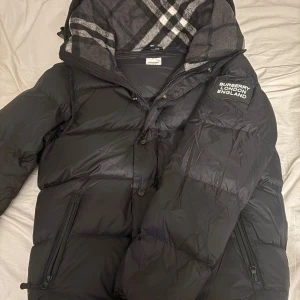 Svart pufferjacka från Burberry  - Säljer en svart pufferjacka från Burberry i storlek L men passar M. Jackan har en stor patch med Burberry London England på ärmen, dragkedjor på fickorna och en huva fodrad med klassiskt rutigt tyg i grått och vitt. Perfekt för kalla dagar och riktigt snygg streetstil.