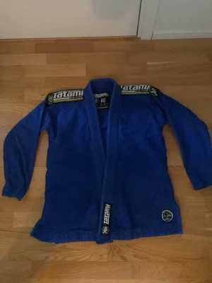Blå BJJ gi från Tatami - Säljer en blå brasiliansk jiu-jitsu gi från Tatami med broderade loggor på axlar och nederkant. Tillverkad i slitstarkt material som passar perfekt för träning och tävling. Klassisk design med förstärkta sömmar och tjock krage.