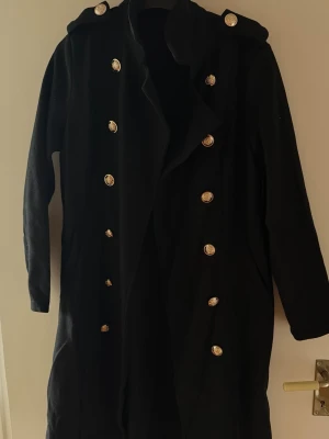 Svart lång kappa med guldknappar - Stilren svart lång kappa med dubbelknäppning och dekorativa guldknappar framtill. Kappan har bred krage och en klassisk, rak passform som ger en snygg siluett. Perfekt för dig som vill ha en elegant och tidlös look till höst och vinter.