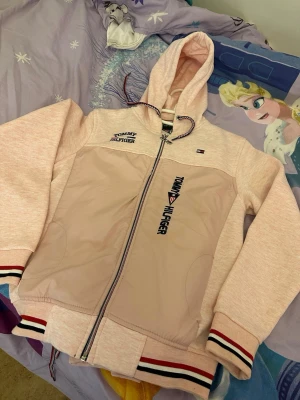 Ljusrosa hoodie  - Ljusrosa hoodie med dragkedja, broderad logga på bröstet och ärmen samt två fickor fram. Huva med dragsko och ribbade muddar med röd, vit och svart rand. Materialmix med mjuk bomull och glansig panel framtill.