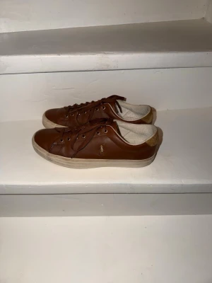 Bruna sneakers från Polo Ralph Lauren - Snygga bruna sneakers i skinn från Polo Ralph Lauren med klassisk logga på sidan. Skorna har rund tå, snörning och vit platt sula. Insidan är fodrad i beige textil och hälen har en kontrastdetalj i mocka. Perfekta till jeans eller chinos. Jag säljer på grund av fel storlek. Priset kan diskuteras. Avhämtning som gäller.
