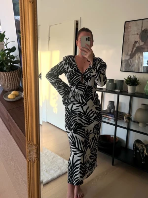 Svartvit långklänning - Snygg långklänning i svart och vitt med stort bladmönster. Klänningen har v-ringning och långa ärmar. Perfekt för dig som vill sticka ut med ett trendigt print och en feminin siluett