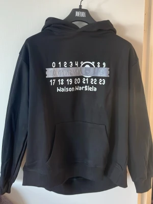 Svart Maison Margiela hoodie - Svart hoodie från Maison Margiela med tryckt logga och siffror på bröstet. Klassisk känguruficka framtill och huva. Tillverkad i mjuk bomull, perfekt för en avslappnad och trendig look.