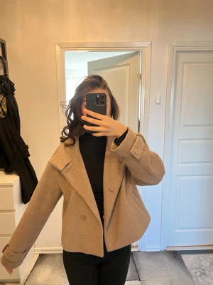 Kort beige ullkappa med bred krage - Snygg kort beige kappa med bred krage och dubbelknäppning framtill. Kappan har en rak passform och uppvikta ärmslut, vilket ger en trendig och stilren look. Perfekt för dig som vill ha en klassisk men modern jacka till hösten eller våren. Kappan har bara använts några gånger vilket betyder att det är i en mycket bra skick. Storleken är 34 och pris kan diskuteras 