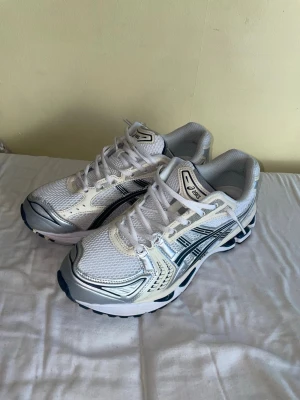 Vita ASICS GEL-1130 sneakers - Säljer ett par vita ASICS GEL-1130 sneakers med detaljer i silver och mörkblått. Skorna har mesh- och syntetmaterial, klassisk sportig design och tydliga ASICS-loggor på sidorna. Perfekta för dig som gillar retrostil och bekväma sneakers.