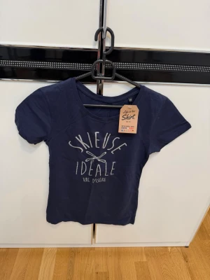 Mörkblå t-shirt med tryck - Mörkblå t-shirt från Life is a Shirt med vitt tryck framtill där det står 'SKIEUSE IDEALE VAL D'ISERE' och två korsade skidor. T-shirten har korta ärmar och en klassisk rund halsringning. 