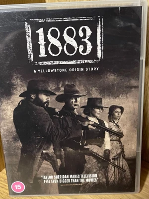 1883: A Yellowstone Origin Story DVD - DVD-box med serien '1883: A Yellowstone Origin Story'. Omslaget har ett svartvitt westernmotiv med cowboys och pionjärer. Serien är skapad av Taylor Sheridan och är en prequel till Yellowstone. Innehåller över två timmar extramaterial. Text och ljud på engelska.