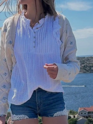 Vit blus med volang och knappar ärmlös  - Supersöt vit blus från Co couture med volangdetaljer på ärmarna, knappar framtill och lätt rynkad struktur. Blusen är  ärmlös  och är gjord i tunn, luftig bomull – perfekt för varma dagar. Passar till både jeans och kjol för en fräsch look. Ok