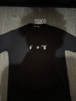 Svart Off-White t-shirt med tryck - Säljer en svart t-shirt från Off-White med ett litet grafiskt tryck på bröstet och en stor, färgstark grafisk design på ryggen i vitt, blått och orange. Klassisk rund hals och korta ärmar. Perfekt för dig som gillar streetwear och vill sticka ut.