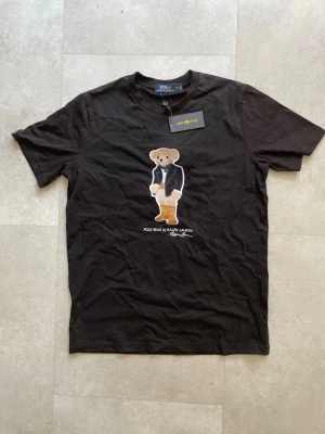 Svart Polo Bear t-shirt från Ralph Lauren - Svart t-shirt från Ralph Lauren med ikoniska Polo Bear-trycket på bröstet. Klassisk passform och rund hals. Tillverkad i mjuk bomull som känns skön mot huden. Perfekt för dig som vill ha en stilren men lekfull look.