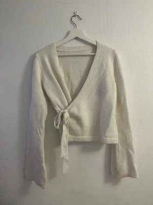 Vit omlottstickad tröja - Säljer en krämvit stickad omlottopp med långa ärmar och knytband i sidan. Tröjan har ribbade muddar vid ärmslut och nederkant, samt en djup v-ringning. Perfekt för lager på lager och enkel att styla till olika outfits.