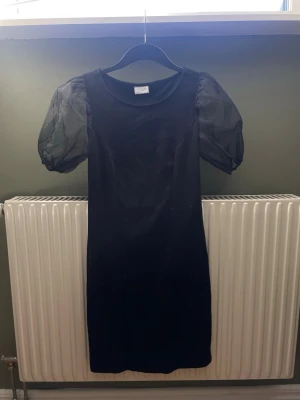Svart klänning med puffärm från Vila - Stilren svart klänning från Vila med transparenta puffärmar i organza och rak passform. Klänningen är knälång och har rund halsringning. Perfekt för dig som vill ha en enkel men trendig look.