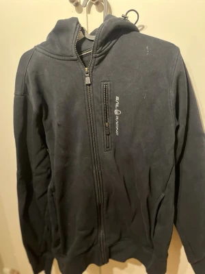 Svart hoodie från Sail Racing - Säljer en svart hoodie från Sail Racing med dragkedja framtill och en bröstficka med dragkedja. Hoodien har broderad logga på bröstet och en klassisk huva. Perfekt för dig som gillar sportig och stilren look.