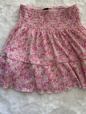 Blommig kortkjol i rosa toner - Supersöt kortkjol från Gina med blommigt mönster i rosa, vitt och grönt. Kjolen har en bred smock i midjan och volangdetalj nedtill för en extra flowy känsla. Perfekt för varma dagar och ger en romantisk vibe till din outfit. Använd ett fåtal gånger men inget som syns!