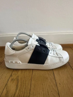 Valentino vita sneakers med svart detalj - Snygga vita sneakers från Valentino med bred svart läderdetalj över mitten och coola nitar på hälen. Skorna har klassisk låg profil, vita snören och diskret logga på plösen. Tillverkade i läder för en lyxig känsla och stilren look.