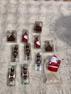 Julgranspynt med nötknäppare och tomtar - Säljer ett set julgranspynt med fyra röda och guldiga nötknäppare, tre röda tomtar, tre guldfärgade hus och en större tomtefigur. Alla dekorationer är i plast eller glas och kommer i sina originalförpackningar. Perfekt för att pynta granen till jul!