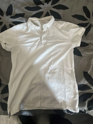 Vit piké t-shirt från Kappahl - Klassisk vit piké t-shirt från Kappahl med korta ärmar och krage. Tröjan har knappar framtill och är tillverkad i mjuk bomull, perfekt för en clean och enkel stil.