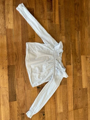 Vit offshoulder blus med volang - Säljer en söt vit offshoulder blus med långa ärmar och volangdetalj vid halsringningen. Blusen har resår vid axlar och ärmslut för en bekväm passform. Perfekt att styla med jeans eller kjol för en trendig look.