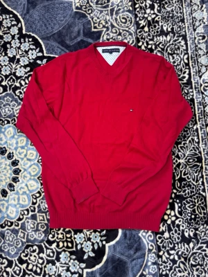 Röd v-ringad tröja Tommy Hilfiger - Snygg röd v-ringad tröja från Tommy Hilfiger med diskret logga på bröstet. Tröjan har långa ärmar, ribbade muddar och är tillverkad i ett mjukt material som känns skönt mot huden. Perfekt för dig som vill ha en klassisk och stilren look.