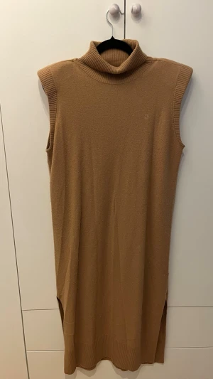 Beige stickad poloklänning - Snygg beige stickad klänning med polokrage och ärmlös design. Klänningen har ribbade detaljer vid krage och nederkant samt små slitsar i sidorna. Perfekt att styla med boots eller sneakers för en trendig look. Defekt bak små hål men syns knappt.