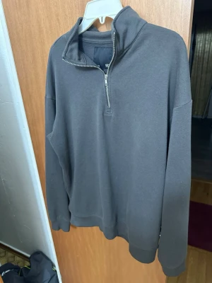 Mörkgrå halvzip tröja - En stilren mörkgrå stickad tröja med halv dragkedja och hög krage. Tröjan har långa ärmar och ribbade muddar vid ärmslut och nederkant. Perfekt för lager på lager och enkel att matcha med olika outfits.