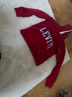 Röd hoodie från Levi's med logga - Snygg röd hoodie från Levi's med stor vit och blå logga på bröstet. Klassisk modell med huva och långa ärmar. Tillverkad i mjukt bomullsmaterial som är skönt att ha på sig. Perfekt för en avslappnad och trendig look.