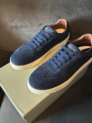 Marinblå sneakers Brunello Cucinelli äldre modell - Stilrena marinblå sneakers från Brunello Cucinelli i mocka med vita sulor och blå skosnören. Insidan har en snygg kontrast i beige läder och loggan syns i guld på plösen. Perfekta för dig som gillar exklusiv och clean design.