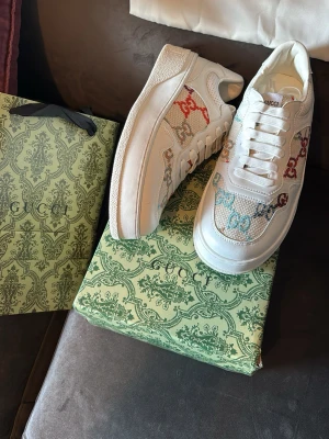 Gucci vita sneakers med färgglada GG - Säljer ett par vita Gucci sneakers med färgglada GG-loggor i rött, blått och beige. Skorna har en chunky sula, vita skosnören och detaljer i mesh och läder. Klassisk låg modell med exklusiv känsla och tydlig Gucci-branding på sulan.