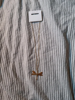 Guldfärgat halsband med trollslända - Snyggt halsband från Edblad i guldfärgad metall med ett stort hänge i form av en stilren trollslända. Kedjan är tunn och elegant, och trollsländan har en modern, blank finish som verkligen sticker ut. Perfekt accessoar för dig som gillar unika detaljer och vill addera lite edge till din stil.