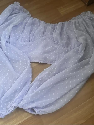 Ljuslila offshoulder blus med puffärm - Supersöt ljuslila offshoulder blus med puffiga långärmar och små prickiga detaljer. Blusen har resår vid axlar och ärmslut för en snygg passform och är gjord i ett tunt, luftigt material som känns perfekt för varmare dagar.