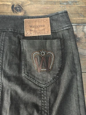 Svart jeanskjol från Weekend Max Mara  - Snygg svart jeanskjol från Weekend med rak passform och dekorativ bakficka med broderad krona och W i brunt och silver. Kjolen har klassisk midja och är tillverkad i slitstarkt denimtyg. Perfekt för en trendig och avslappnad look.