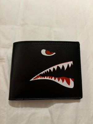 Svart plånbok med hajmotiv - Snygg svart plånbok i skinn med ett coolt hajmotiv på framsidan i vitt och rött. Plånboken har en klassisk fyrkantig form och är perfekt för dig som vill ha en unik accessoar med streetwear-vibe.