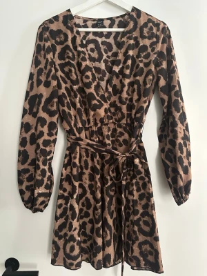 Leopardmönstrad omlottklänning Shein - Kort leopardmönstrad omlottklänning från shein, med långa ärmar och knytband i midjan. Klänningen har v-ringning och är tillverkad i ett mjukt, lätt material som ger en snygg siluett. Perfekt för dig som vill sticka ut med ett djurmönster.