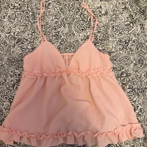 Ljusrosa volanglinne med tunna axelband - Supersött ljusrosa linne med tunna axelband och volangdetaljer både upptill och nedtill. Toppen har en lätt v-ringning och är luftig i modellen, perfekt för varma dagar.