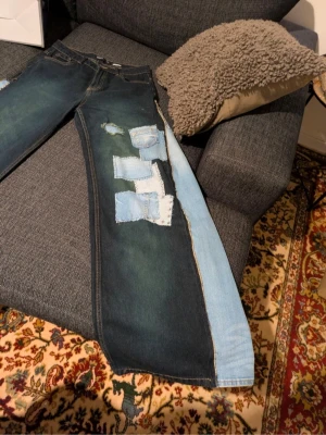 Unika patchade jeans med breda ben - Säljer ett par mörkblå jeans med breda ben och ljusblå patchar på ena sidan. Jeansen har flera handsydda detaljer och kontrasterande tygbitar som ger ett coolt och kreativt uttryck. Materialet är denim och modellen är loose fit.