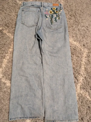 Ljusblå jeans med broderade blommor - Unika ljusblå jeans med raka ben och broderade blommor i gult, vitt och rosa på bakfickan. Jeansen har hög midja och klassisk femficksdesign. Perfekta för dig som vill sticka ut med en kreativ och personlig stil.
