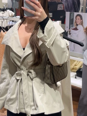 Beige trenchcoat med knytskärp - Stilren beige trenchcoat med bred krage och knytskärp i midjan. Jackan har stora knappar framtill och dekorativa remmar vid ärmsluten. Tillverkad i ett slätt tyg som ger en klassisk look. Perfekt för dig som vill ha en tidlös och trendig jacka.